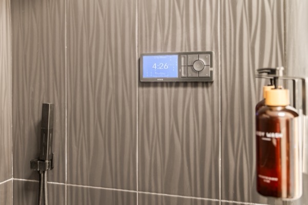 A digital shower control panel and a handheld shower on textured tile, next to a bottle of soap