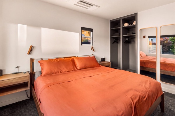 Primary bedroom from the door: a king bed in rust-colored linens against a white wall with reading sconces, an open closet and two tall mirrors