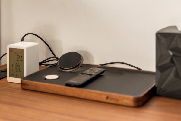 A wood charging tray on the nightstand with a wireless charger and a TV remote, beside an air-quality monitor