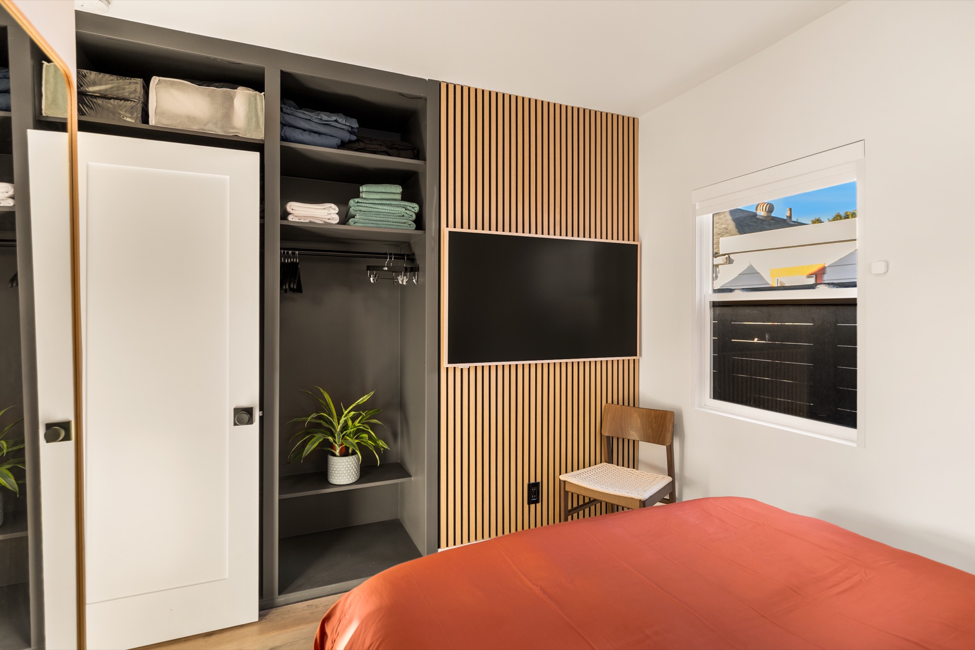 Second bedroom with a closet of open shelves and folded linens, a TV on a light wood slat wall, and the foot of the bed in rust-colored linens
