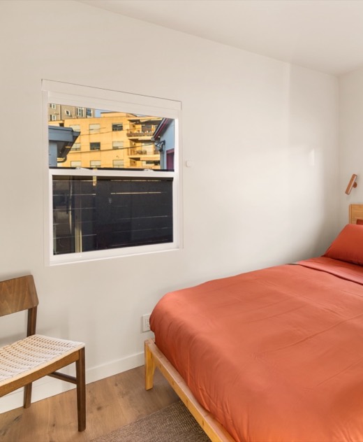 Second bedroom with a queen bed in rust-colored linens on a light wood frame, a woven-seat chair and a window in white walls