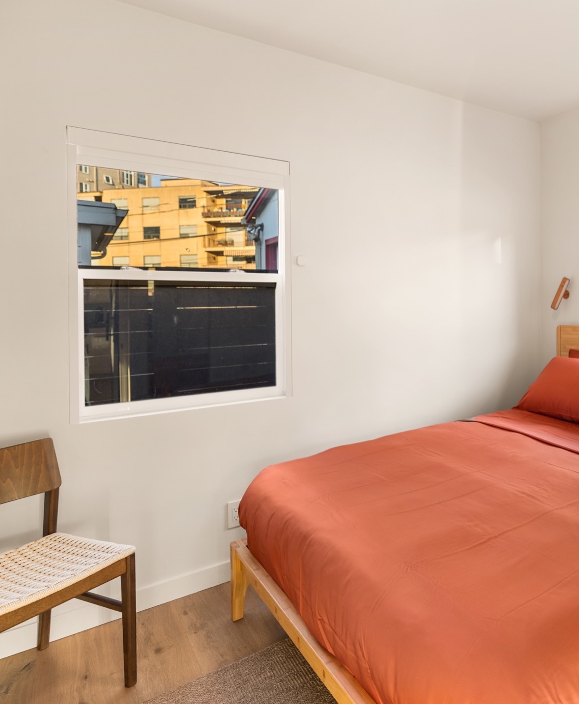 Second bedroom with a queen bed in rust-colored linens on a light wood frame, a woven-seat chair and a window in white walls