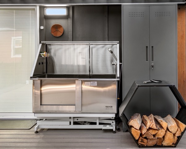 A stainless dog-wash tub with a handheld sprayer on the back deck, beside a storage cabinet and a firewood rack