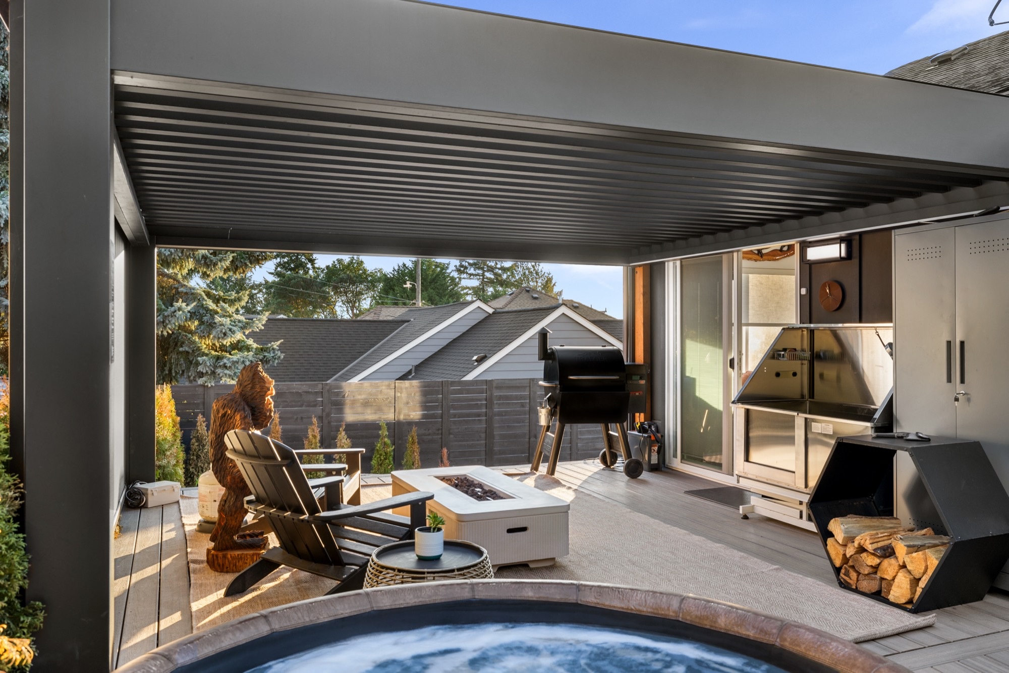 The covered back deck under a louvered pergola: an Adirondack chair, a gas fire pit, a grill and a carved wooden bear, with the rim of the Softub in the foreground.