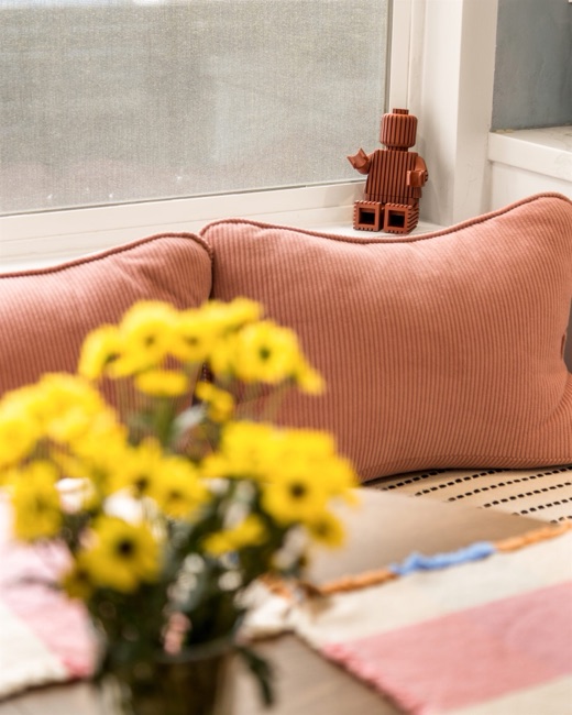 A terracotta toy figure sitting on the windowsill behind the coral cushions of the window bench, with yellow flowers in the foreground