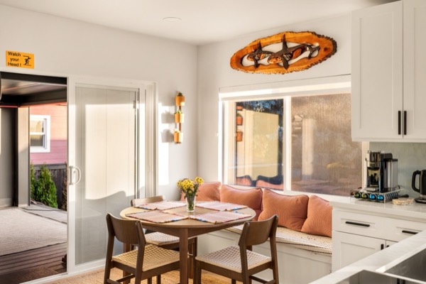 Dining nook with a round wood table, woven-seat chairs and a cushioned window bench lined with coral pillows