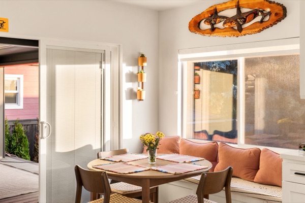 Sunlit dining nook with a round wood table, woven-seat chairs, coral pillows on the window bench and a carved wooden orca panel above, beside the sliding door to the deck