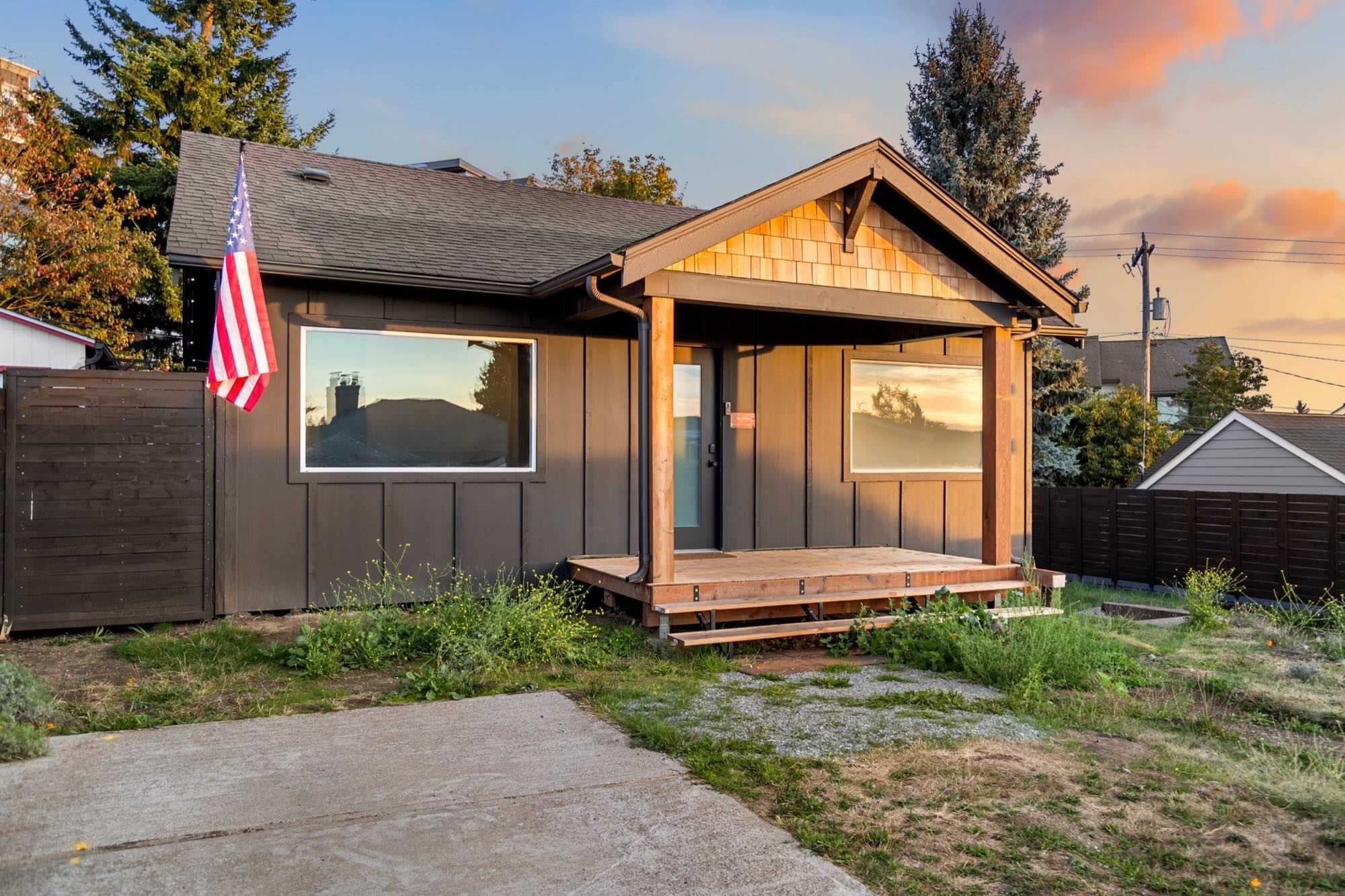 The house at golden hour: a covered front porch under a shingled gable, big picture windows and tall evergreens behind.