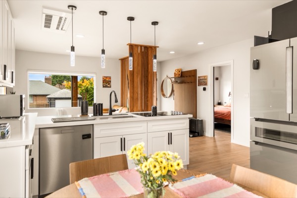 The kitchen seen from the dining table: white cabinets, a dishwasher, a stainless fridge and pendant lights over the island
